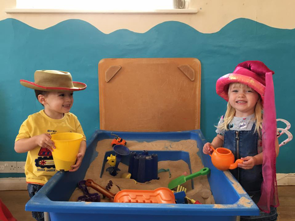 Two children stand either side of an indoor sandpit