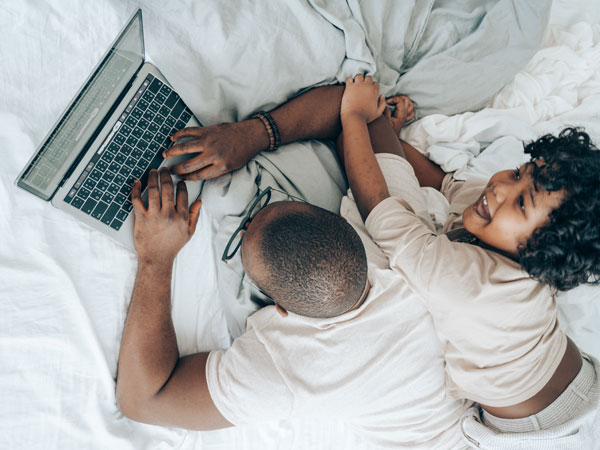 A man lies on a bed using a laptop whilst a child distracts him