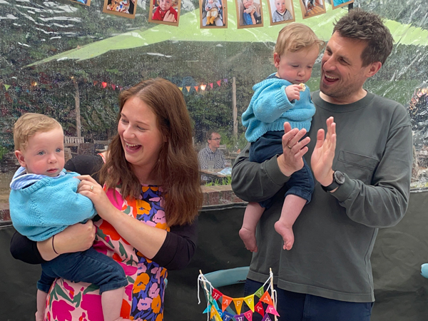 A mother and father at a birthday celebration each holding a twin baby